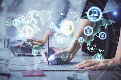 Digital composite image showing people working on financial documents and laptops with a holographic world map overlay featuring currency symbols such as the dollar, euro, yen, and pound, representing global finance and international currency exchange.